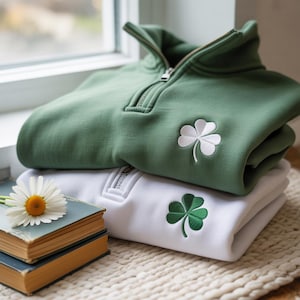 May include: Two folded sweatshirts, one green and one white, are stacked. Each has a quarter-zip neckline and a shamrock emblem. A daisy rests on a stack of old books in the foreground. The sweatshirts are made of a soft material.