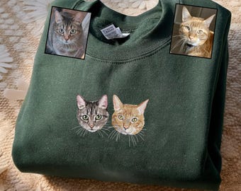 May include: Dark green sweatshirt featuring embroidered cat faces. Two embroidered cat faces are at the bottom, with two framed cat photos at the top. The sweatshirt is a solid color, with a crew neck and long sleeves. The cat faces are detailed with whiskers and realistic features.