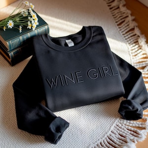 Puede incluir: Sudadera negra con la inscripción bordada "WINE GIRL". La parte superior de manga larga se muestra sobre una alfombra blanca y beige estampada, con una pila de libros y margaritas en el fondo.