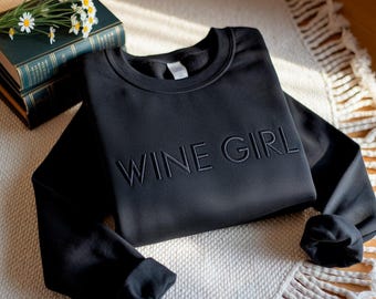 Peut inclure: Sweat-shirt noir avec l'inscription brodée "WINE GIRL". Le haut à manches longues est présenté sur un tapis blanc et beige à motifs, avec une pile de livres et des marguerites en arrière-plan.