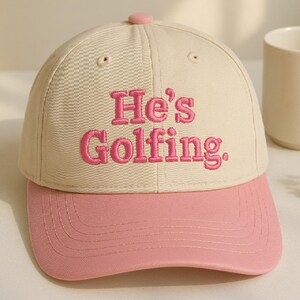 May include: A beige and pink baseball cap with the embroidered text "He's Golfing." The cap features a pink brim and button, with the text in a matching pink color. The hat is a classic six-panel design.