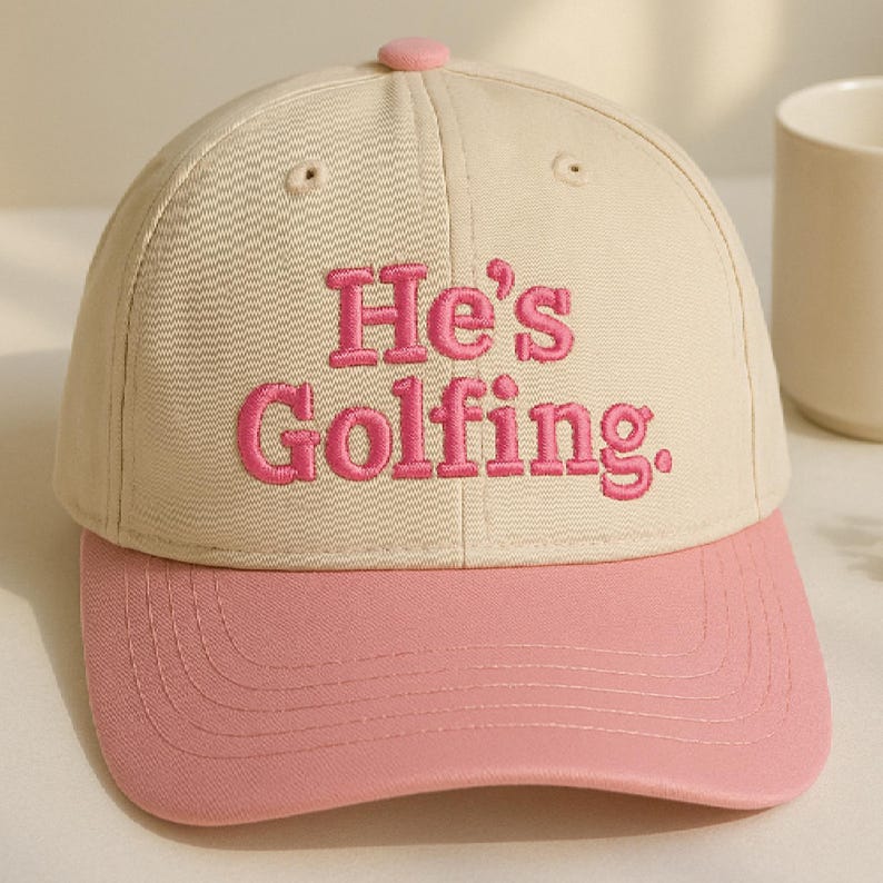 May include: A beige and pink baseball cap with the embroidered text "He's Golfing." The cap features a pink brim and button, with the text in a matching pink color. The hat is a classic six-panel design.