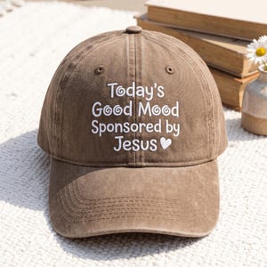 Könnte beinhalten: Eine braune Baseballkappe mit weißem Sticktext, der Today's Good Mood Sponsored by Jesus lautet, ergänzt durch ein kleines Herzsymbol. Die Kappe verfügt über eine strukturierte Krone, einen gebogenen Schirm und Belüftungsösen.