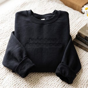 Puede incluir: Una sudadera negra de cuello redondo con el texto bordado "dancing through the lightning strikes" en hilo negro a juego en el pecho. La prenda tiene mangas largas con puños acanalados y un cuello redondo acanalado, presentada de forma sencilla.