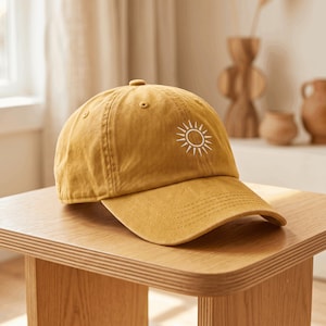 May include: A mustard yellow baseball cap featuring a white embroidered sun icon on the front panel. The cap has a curved brim, a button on the top crown, and stitched eyelets for ventilation, constructed from a solid-colored fabric material.