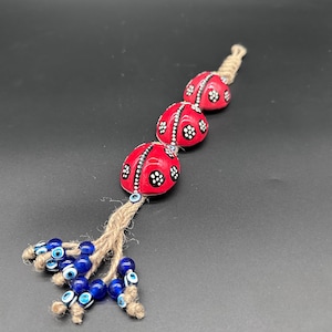 May include: A decorative hanging ornament with three red, ladybug-shaped beads, each with black and white patterns. The beads are strung on a natural twine cord, with a cluster of blue and white evil eye beads at the bottom.