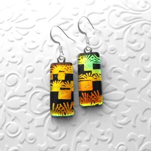 May include: A pair of rectangular dangle earrings with a black background and a geometric pattern in shades of gold and green. The earrings have silver-colored fish hook ear wires.