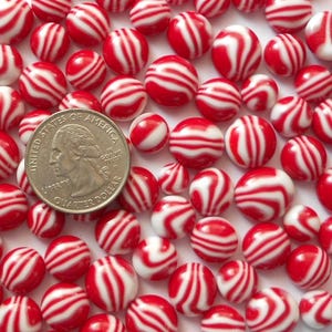May include: A collection of red and white striped beads, resembling peppermint candies, scattered across a surface. A United States quarter coin is included for size comparison. The beads are round and have a glossy finish.