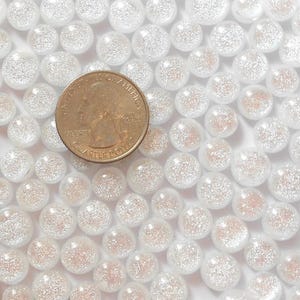 May include: A collection of small, clear, round glass beads with a glittery, iridescent interior. A U.S. quarter coin is included for size comparison. The beads are scattered across a white surface, creating a sparkling effect. These beads are suitable for crafts and jewelry making.