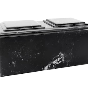 May include: A black marble cremation urn with two separate compartments. The urn has a smooth, polished finish and a rectangular shape.