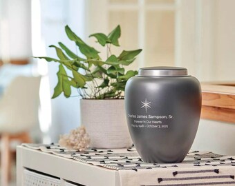 May include: A gray cremation urn with a silver lid. The urn has a star design and is engraved with the text "Charles James Sampson, Sr. Forever In Our Hearts May 19, 1948 - October 3, 2021".