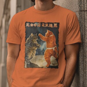 May include: Orange t-shirt with a retro graphic of two cats in a fighting stance. The illustration shows one cat with a white belt. The shirt design includes Chinese characters at the top, giving it a vintage aesthetic.