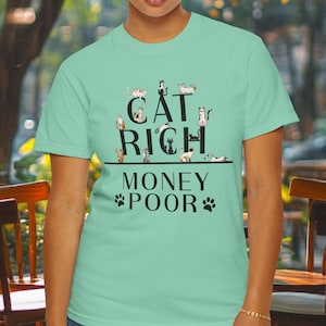 May include: A light green t-shirt with the text "CAT RICH MONEY POOR" in black. The word "CAT" has several cat illustrations above it. Paw prints are on either side of the word "POOR".