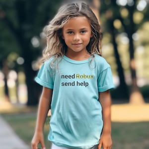 May include: A young person wearing a light blue t-shirt with the text "need Robux send help" in black and yellow lettering. The person is standing in a park with green grass and trees in the background.