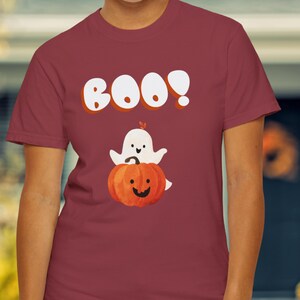Peut inclure: T-shirt bordeaux avec un motif d'Halloween. Le motif comprend le mot "BOO!" en blanc avec un contour orange, un fantôme blanc et une citrouille orange avec un visage souriant.
