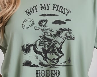 May include: A sage green t-shirt with a black graphic of a cowgirl riding a horse. The graphic includes the text "Not My First Rodeo".