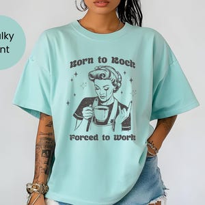 May include: A light blue t-shirt with a retro-style illustration of a woman holding a cup of coffee. The text "Born to Rock, Forced to Work" is printed on the shirt.
