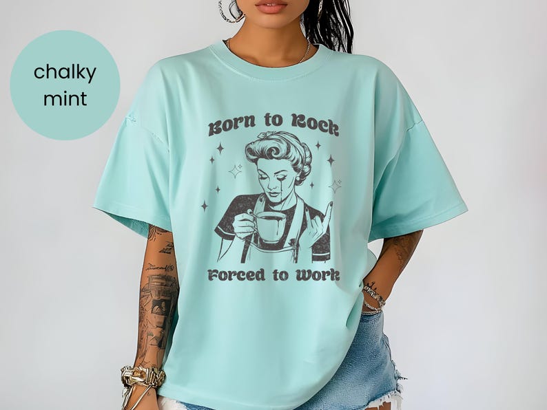 May include: A light blue t-shirt with a retro-style illustration of a woman holding a cup of coffee. The text "Born to Rock, Forced to Work" is printed on the shirt.