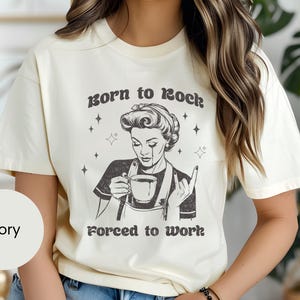 May include: Ivory t-shirt with a black and white illustration of a woman holding a cup of coffee. The text "Born to rock, forced to work" is printed on the shirt.