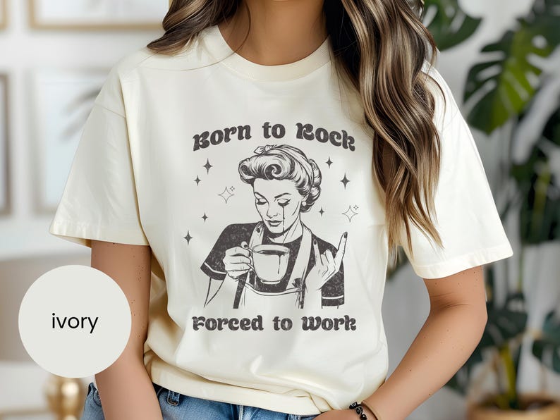May include: Ivory t-shirt with a black and white illustration of a woman holding a cup of coffee. The text "Born to rock, forced to work" is printed on the shirt.