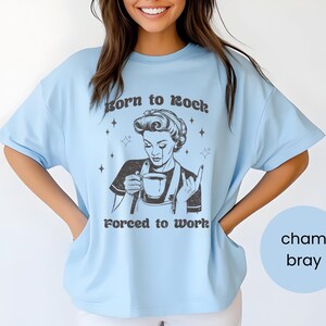 May include: Light blue t-shirt with a retro-style illustration of a woman holding a cup of coffee and making a rock and roll hand gesture. The text on the shirt reads "Born to Rock, Forced to Work."