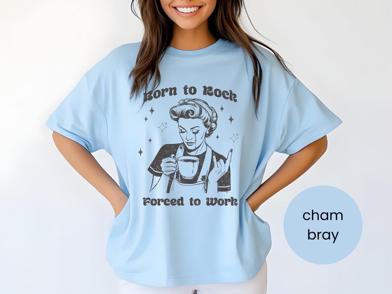 May include: Light blue t-shirt with a retro-style illustration of a woman holding a cup of coffee and making a rock and roll hand gesture. The text on the shirt reads "Born to Rock, Forced to Work."