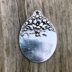May include: A silver oval pendant with a raised floral design. The pendant has a smooth, reflective surface and a hole at the top for hanging.