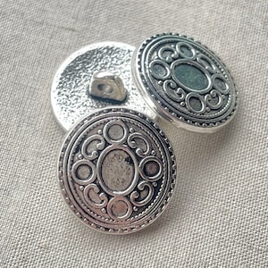 May include: Two silver-toned buttons with an ornate design. The buttons have a raised circular pattern with a smaller circle in the center.