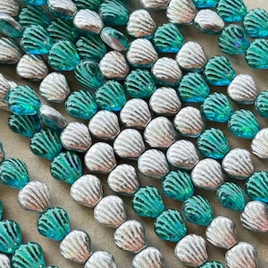 May include: An overhead shot displays a collection of seashell-shaped beads. The beads alternate between silver and teal, with a textured, ridged surface. These decorative beads are ideal for jewelry making or craft projects.