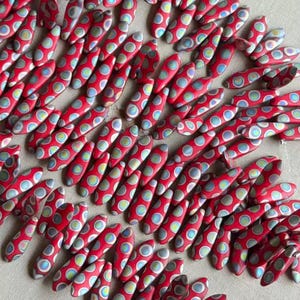 May include: A collection of red, oblong beads with a polka dot pattern in shades of blue, green, and yellow. The beads are arranged closely together, creating a textured surface. These beads could be used for jewelry making or other craft projects.