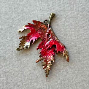 May include: A decorative leaf pendant with a red and gold finish. The metal leaf has a detailed design with a loop at the top for hanging. The pendant is approximately 2 inches long.