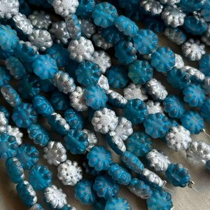 May include: Close-up of strands of beads in shades of blue and silver. The beads are shaped like flowers and are strung together. The beads have a textured surface and a metallic sheen. The beads are arranged in rows.