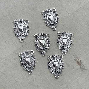 May include: Six silver-toned metal charms with a heart design. Each charm has a detailed ornate border and a heart with a crown on top. The charms have three loops for attaching to jewelry or other crafts.