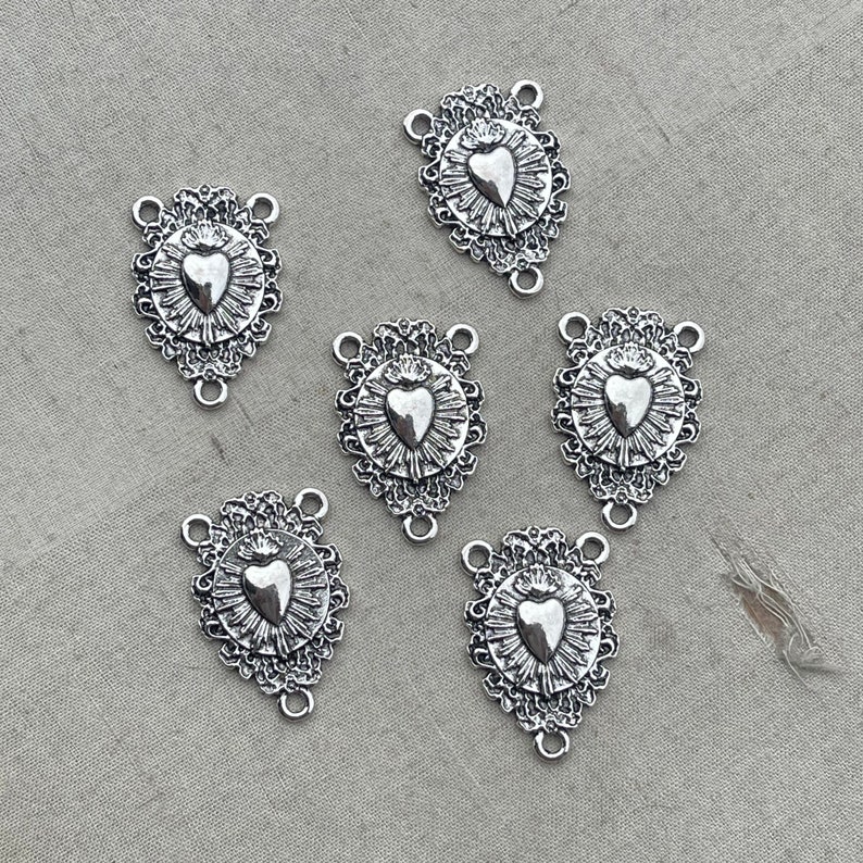 May include: Six silver-toned metal charms with a heart design. Each charm has a detailed ornate border and a heart with a crown on top. The charms have three loops for attaching to jewelry or other crafts.