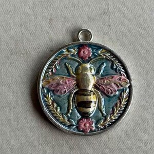 May include: A circular metal pendant featuring a central embossed bee with gold and black stripes and pink-tinted wings. The background is a dark teal enamel with a raised floral border, including small pink flowers at the top and bottom.