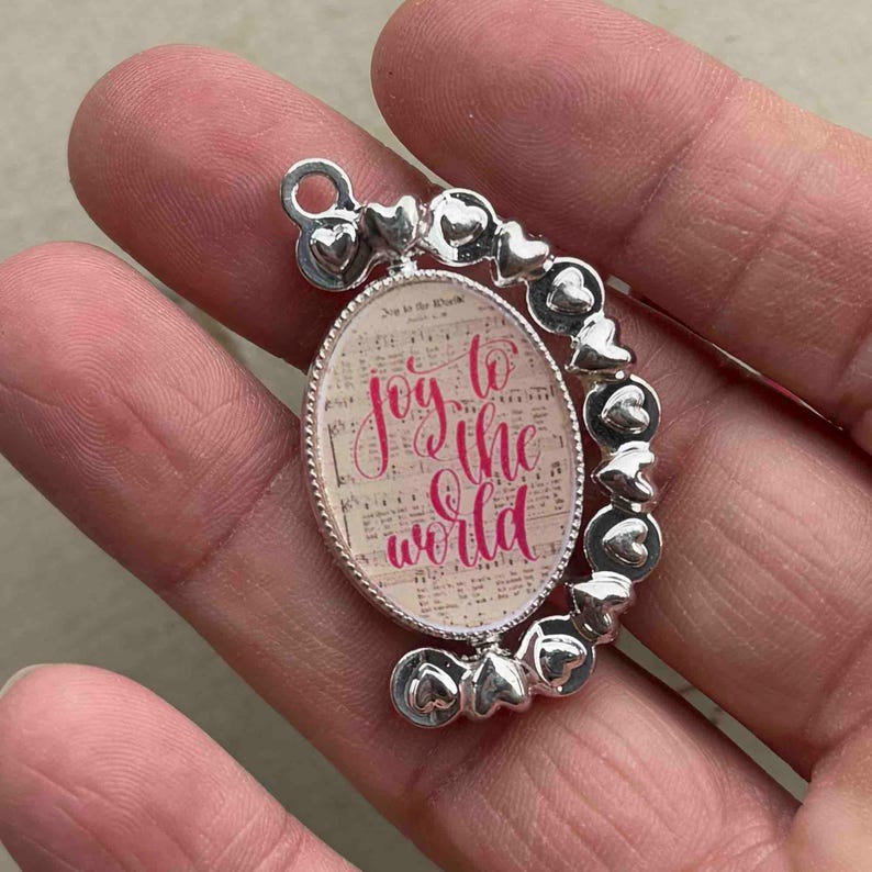May include: Silver-toned pendant with an oval center featuring the words "Joy to the World" in pink script over a musical score background. The pendant is surrounded by a silver frame and heart-shaped accents.