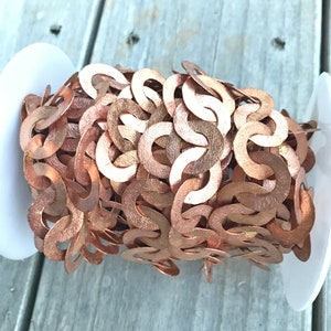 May include: A spool of copper-colored chain. The chain is made of interlocking, textured, crescent-shaped links. The chain is wound around a white plastic spool, set against a weathered wooden surface.