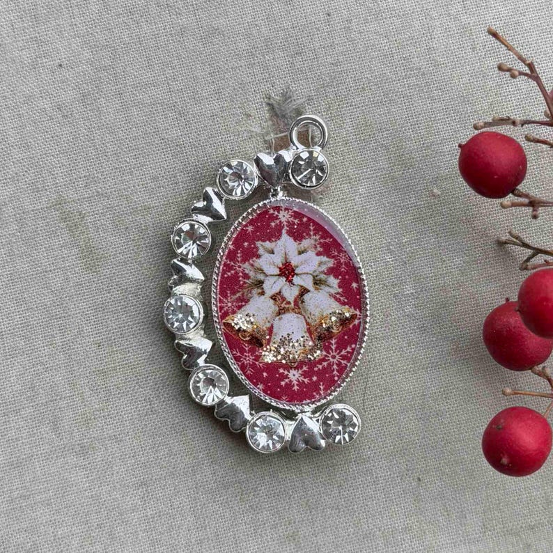May include: Oval Christmas ornament with a red background, white poinsettia, and gold bells. The ornament is surrounded by a silver frame with clear crystals. A sprig of red berries is in the background.