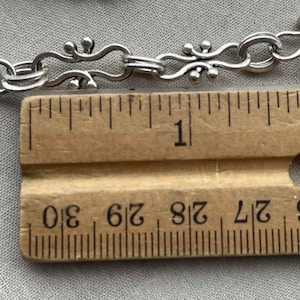 Puede incluir: Primer plano de un collar de cadena de plata con un cierre decorativo. La cadena se mide contra una regla de madera, mostrando que tiene poco m&aacute;s de 1 pulgada de largo.