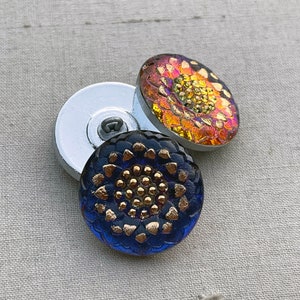 May include: Two glass buttons with a floral design. One button is blue with gold accents and the other is orange with yellow accents.