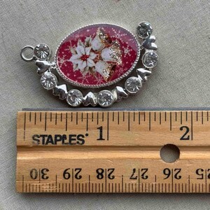 May include: A silver-toned Christmas pendant with a red oval center featuring a poinsettia design and clear crystals. The pendant measures approximately 2 inches in length.