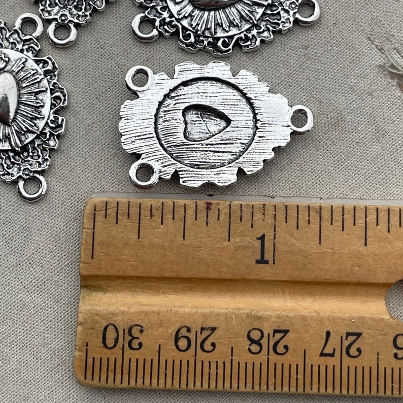 May include: Silver-toned metal charm with a heart design and three jump rings. The charm is approximately 1 inch in diameter and is measured against a ruler.