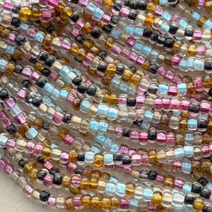 May include: A close-up of a variety of seed beads in different colors, including clear, gold, pink, and blue. The beads are arranged in a random pattern and are all approximately the same size.