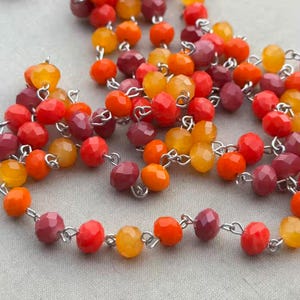 May include: A close-up of a necklace with faceted beads in shades of red, orange, and yellow. The beads are strung together with silver-colored metal links. The necklace is displayed on a light gray surface.