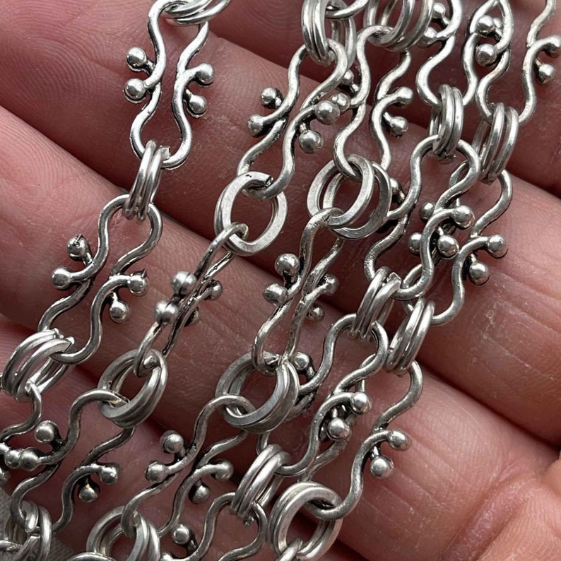 Puede incluir: Primer plano de un collar de cadena de plata con eslabones intrincados y ornamentados. La cadena presenta un patr&oacute;n repetitivo de peque&ntilde;as cuentas redondas y formas delicadas y curvadas.