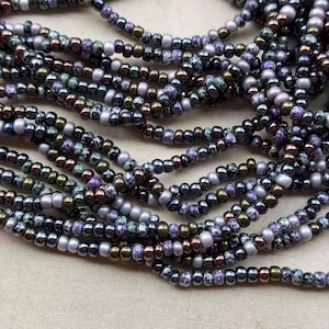 May include: Close-up of multiple strands of beaded necklaces. The beads are a mix of colors, including black, silver, purple, and bronze. The beads are small and round, and the necklaces are layered together.
