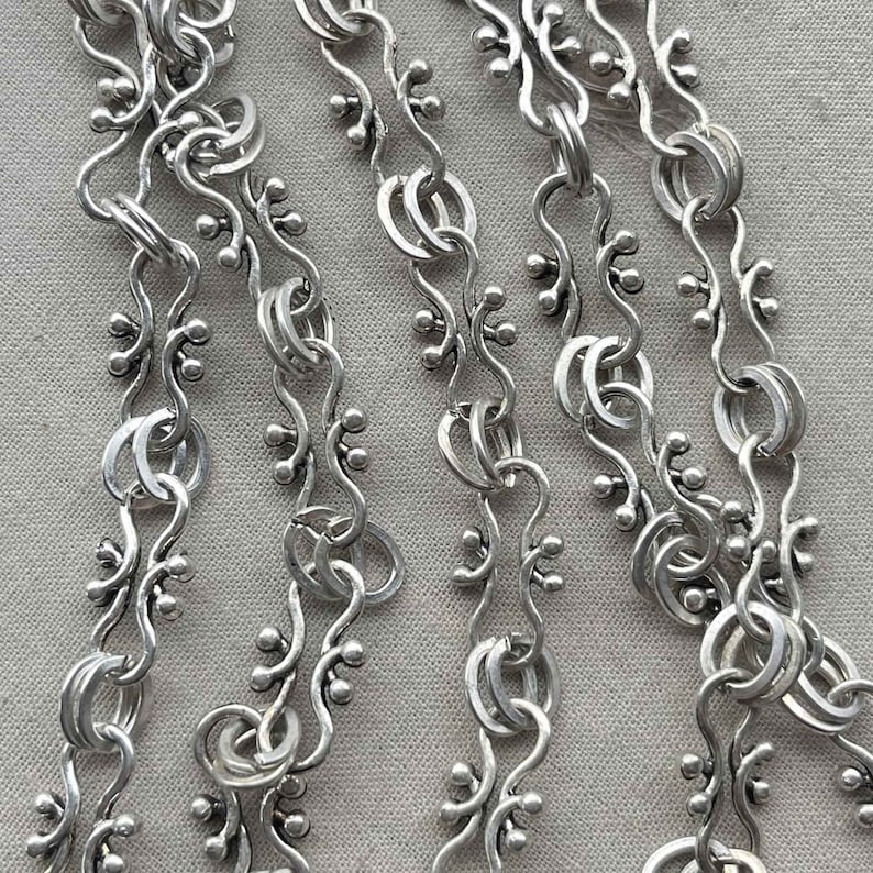 Puede incluir: Primer plano de un collar de cadena de plata con un dise&ntilde;o delicado e intrincado. La cadena presenta un patr&oacute;n repetitivo de peque&ntilde;as cuentas redondas y eslabones curvos.