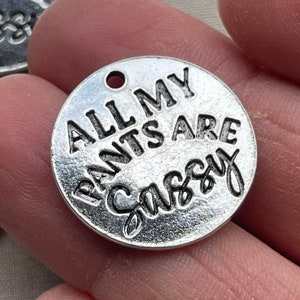 May include: A round, silver-toned charm with the phrase "ALL MY PANTS ARE SASSY" in bold, black lettering. The charm has a small hole at the top for attaching to a necklace or bracelet. The charm is held in a hand.