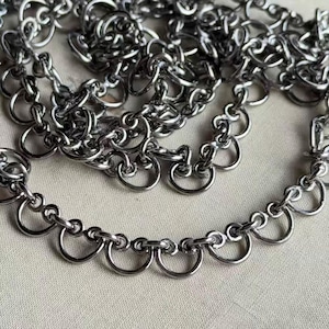 May include: Close-up of a silver-colored chain necklace with a unique design. The necklace features a series of small, connected rings and a decorative, scalloped edge. The metal has a shiny, reflective quality, and the design is intricate.