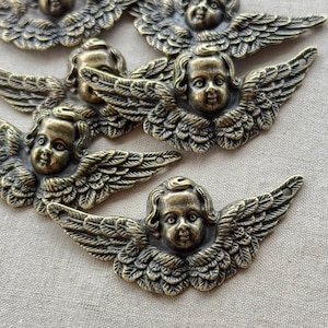 May include: Close-up of several antique brass-toned angel embellishments. Each angel has detailed wings and a serene facial expression. The angels are arranged on a neutral-colored fabric background.